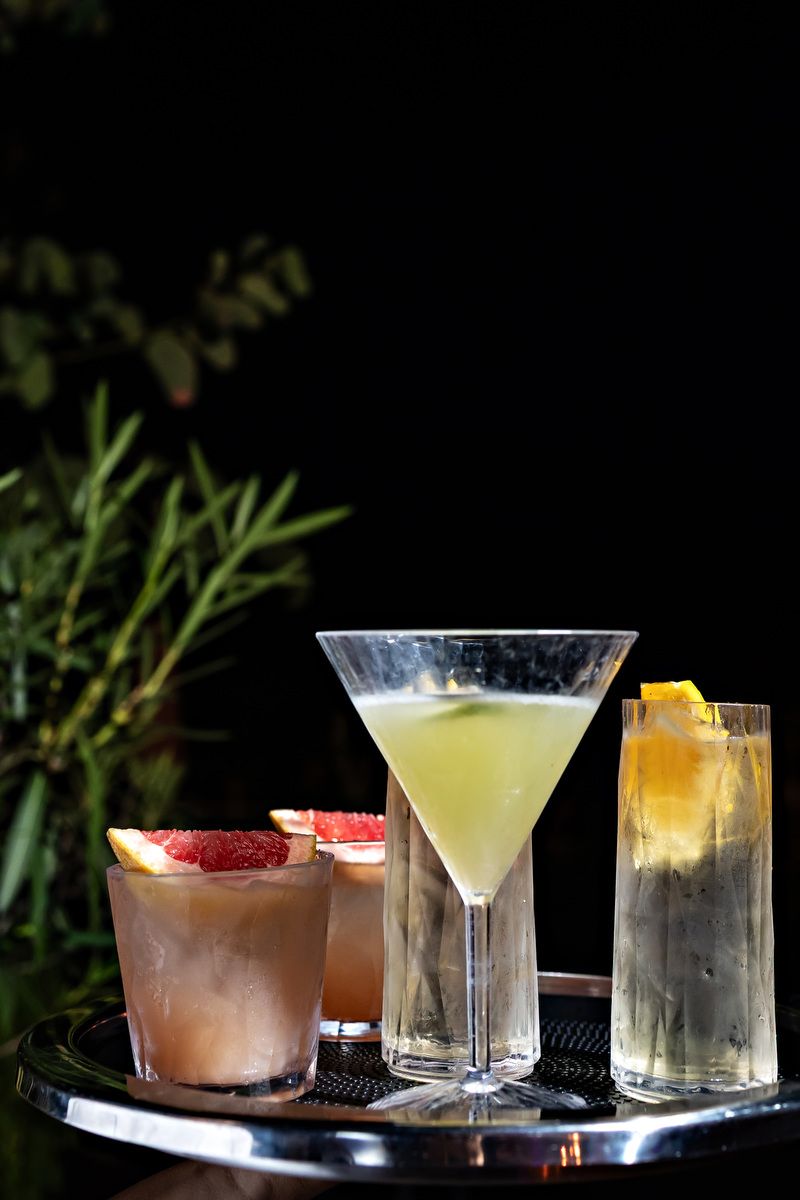 Three elegant cocktails on a silver tray, including a martini and garnished drinks, set against a dark background.