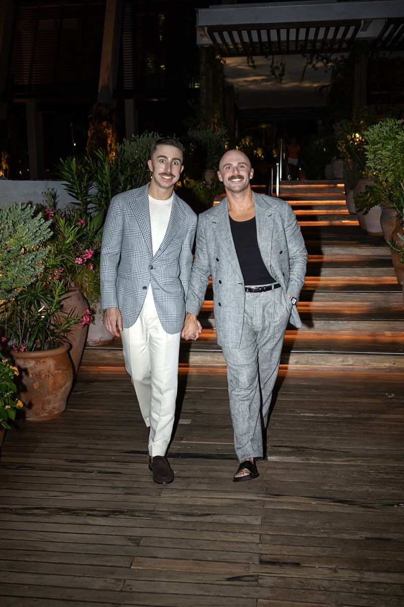 Two stylish men in matching grey suits walk confidently on a wooden deck at night.