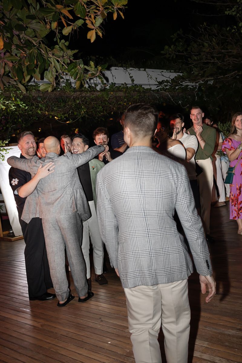 Guests mingling and embracing at an outdoor evening wedding reception on a wooden deck surrounded by lush greenery.
