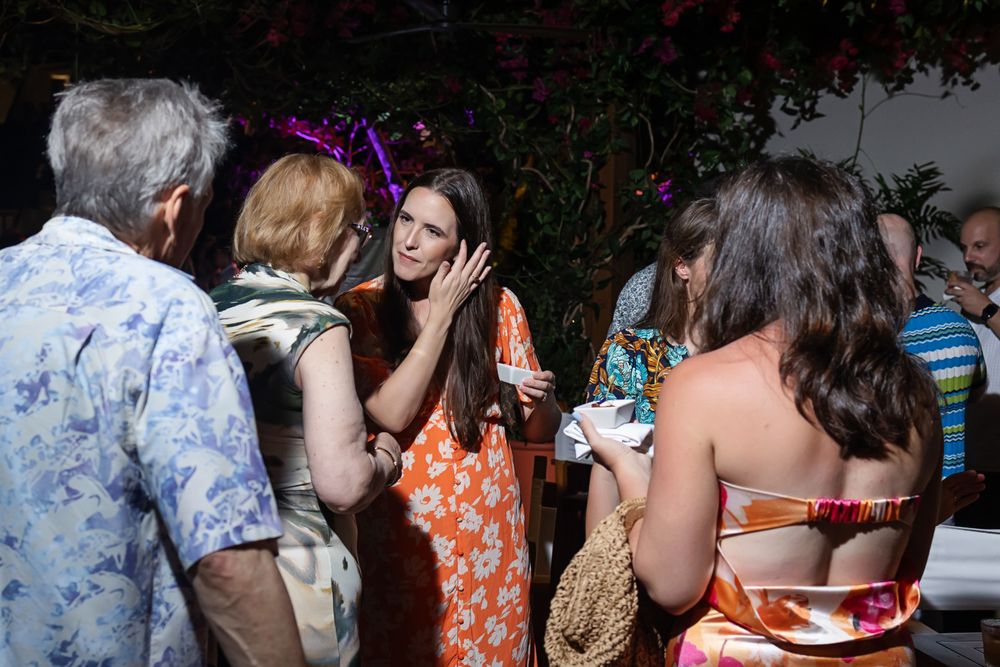 Group of people socializing at an outdoor evening party with colorful lighting and tropical decor.