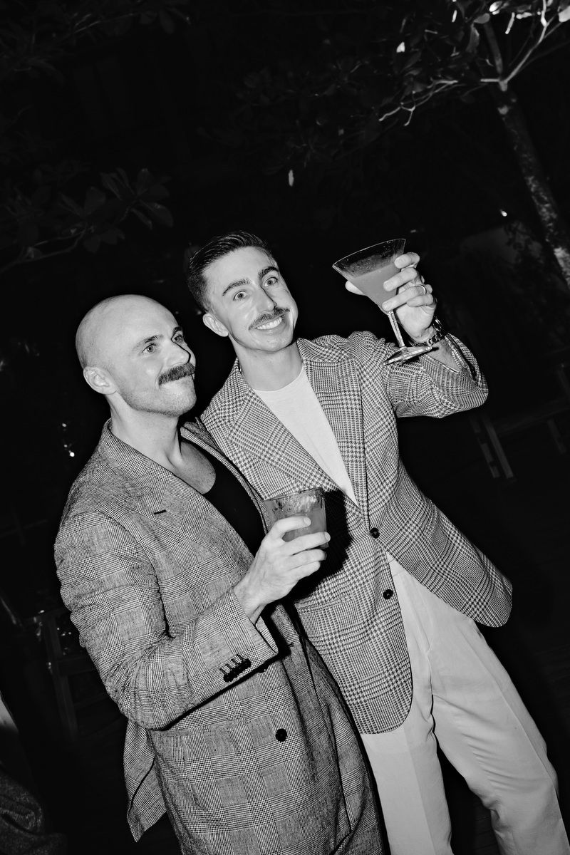 Two men in blazers toast with cocktails at an outdoor evening event in black and white photo.