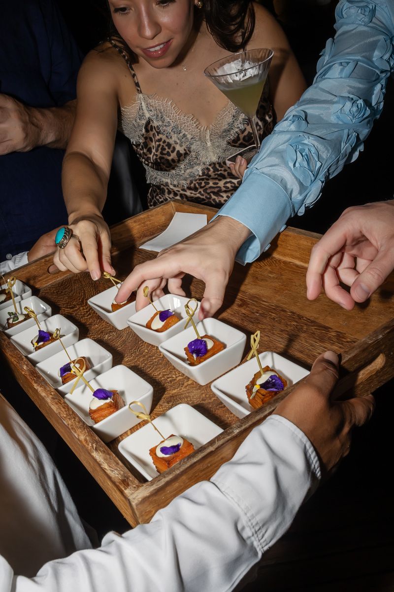Guests reaching for elegant canapés on a wooden tray at a upscale cocktail party event.