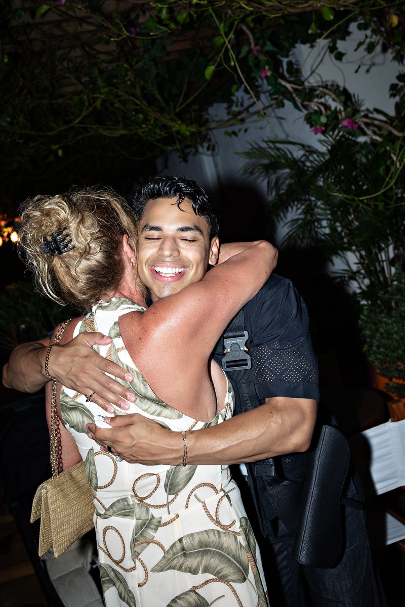 Two people sharing a warm hug at an outdoor evening event, man smiling joyfully, surrounded by tropical greenery.