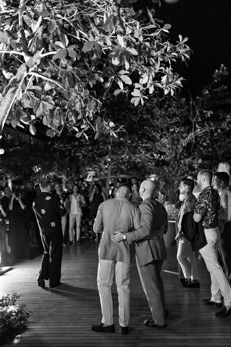 Two men embrace on a wooden dance floor surrounded by lush trees at an outdoor evening event.