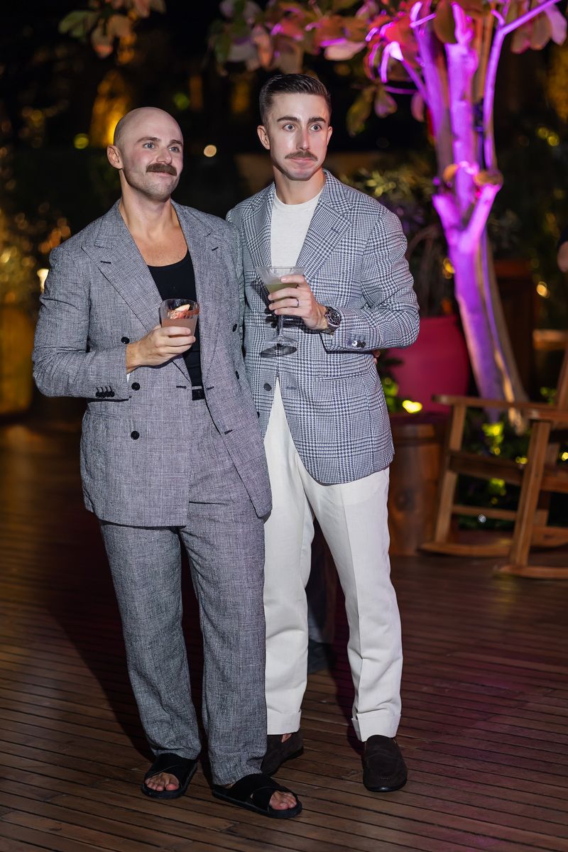 Two men in gray blazers holding cocktails at an outdoor evening event with purple lighting.
