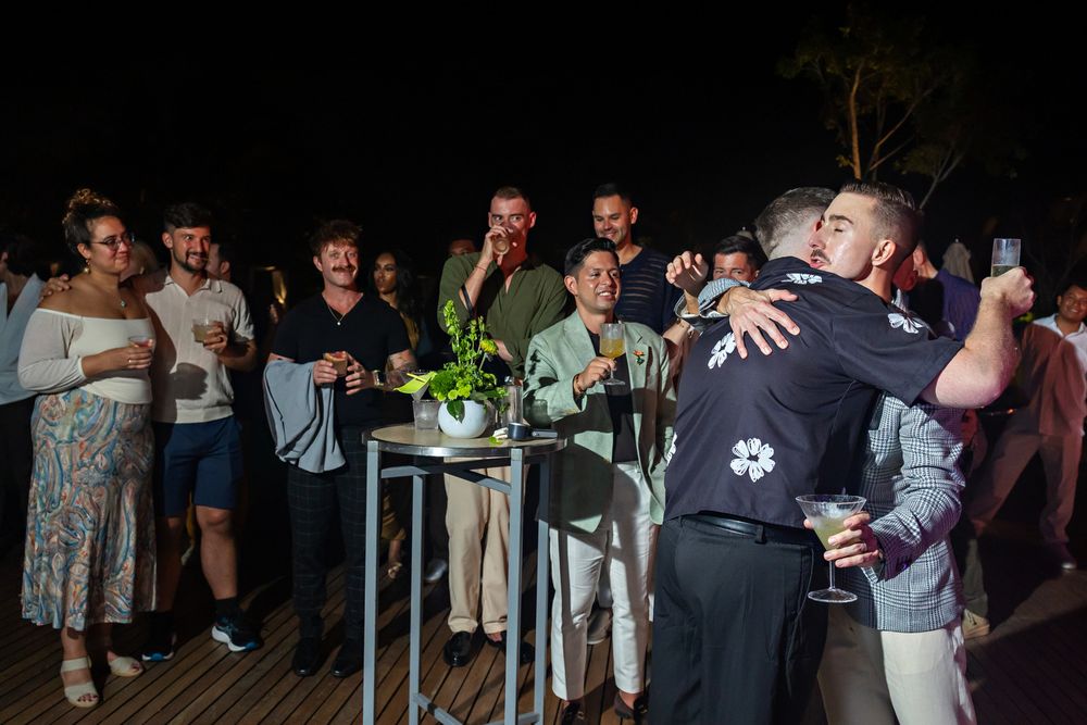 Two men embrace at an outdoor evening celebration while guests socialize with drinks on a deck.