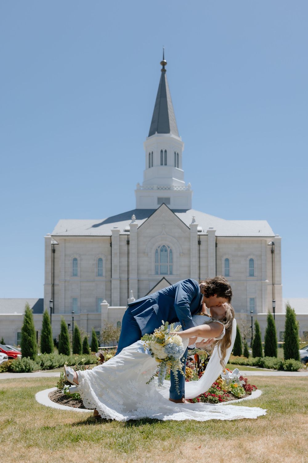 Bride and groom share a romantic dip kiss in front of a white LDS temple with a tall steeple on a sunny day.