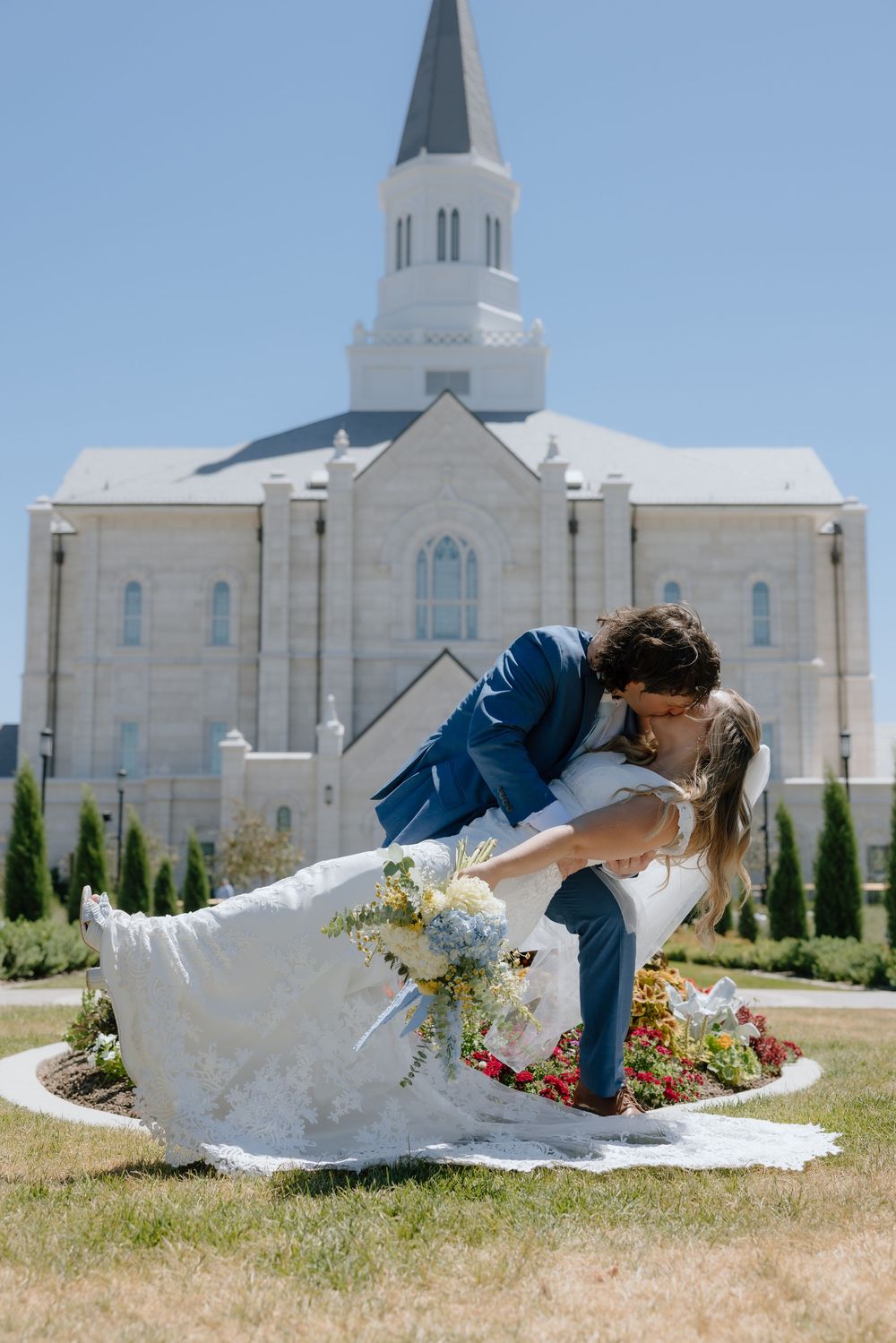 Groom dipping bride in a romantic kiss in front of a white LDS temple with colorful gardens.