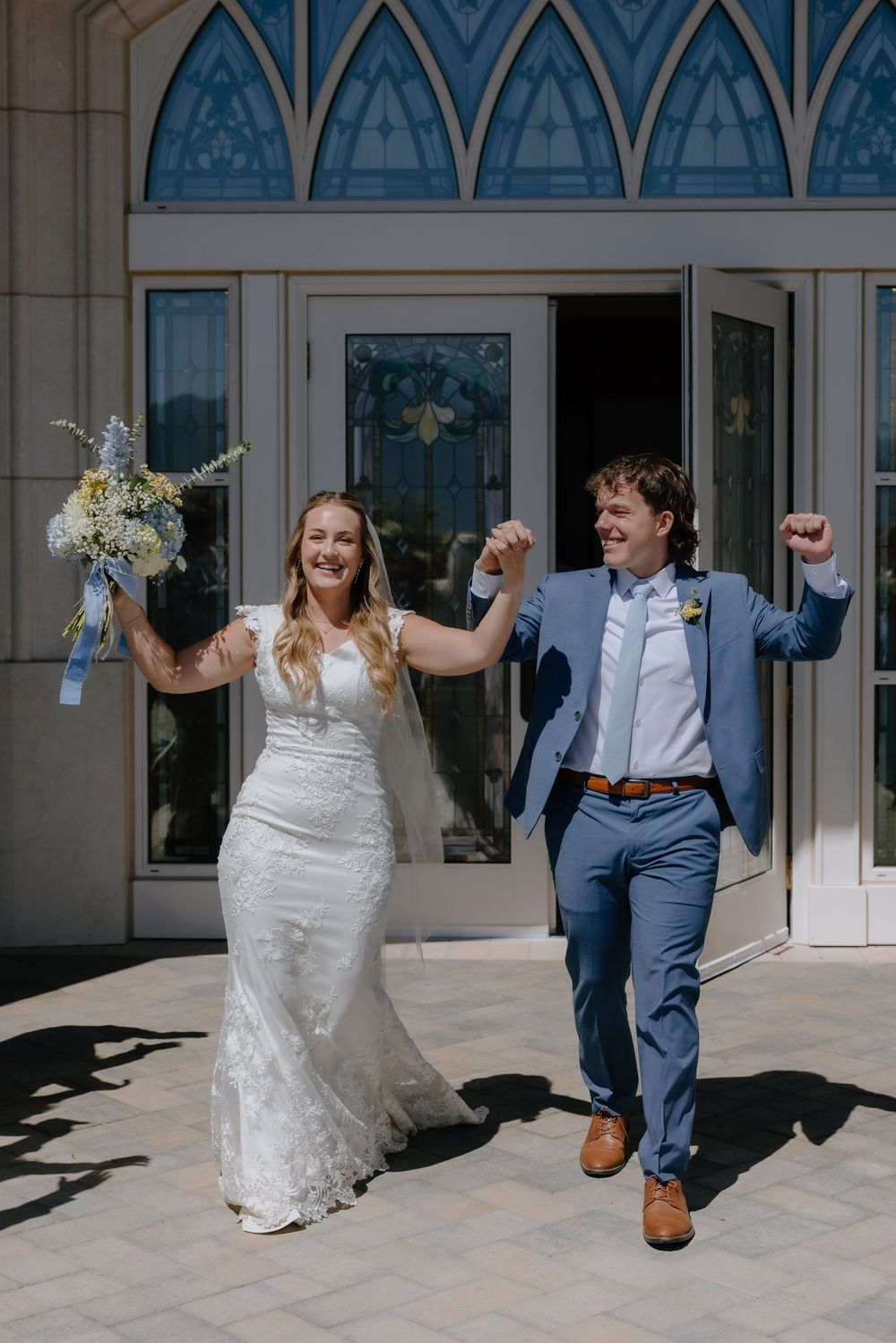 Joyful newlyweds celebrate exiting church, bride in lace gown holding bouquet, groom in blue suit raising fists.
