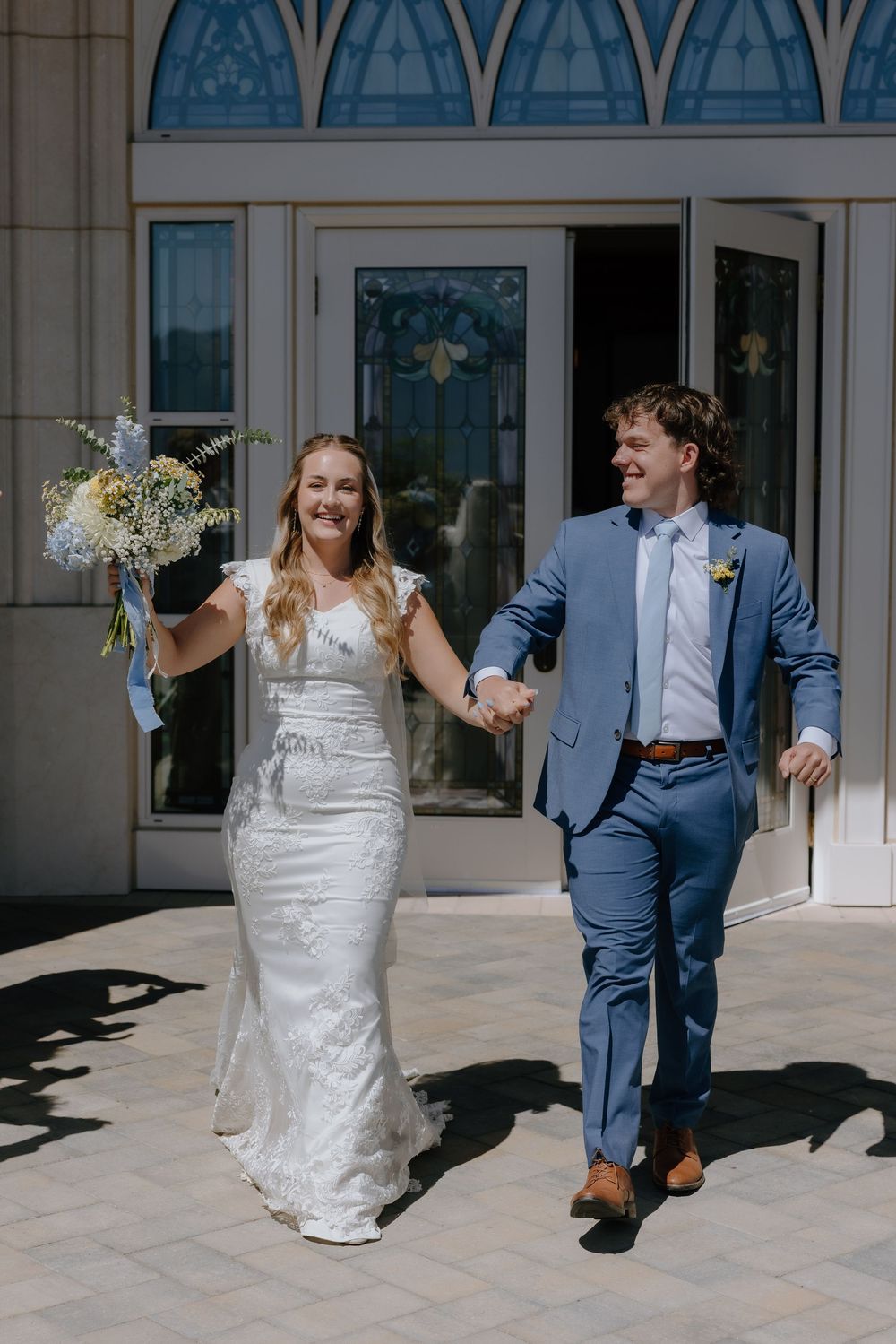 Joyful newlyweds exit church, bride in white gown holding bouquet, groom in blue suit, holding hands.