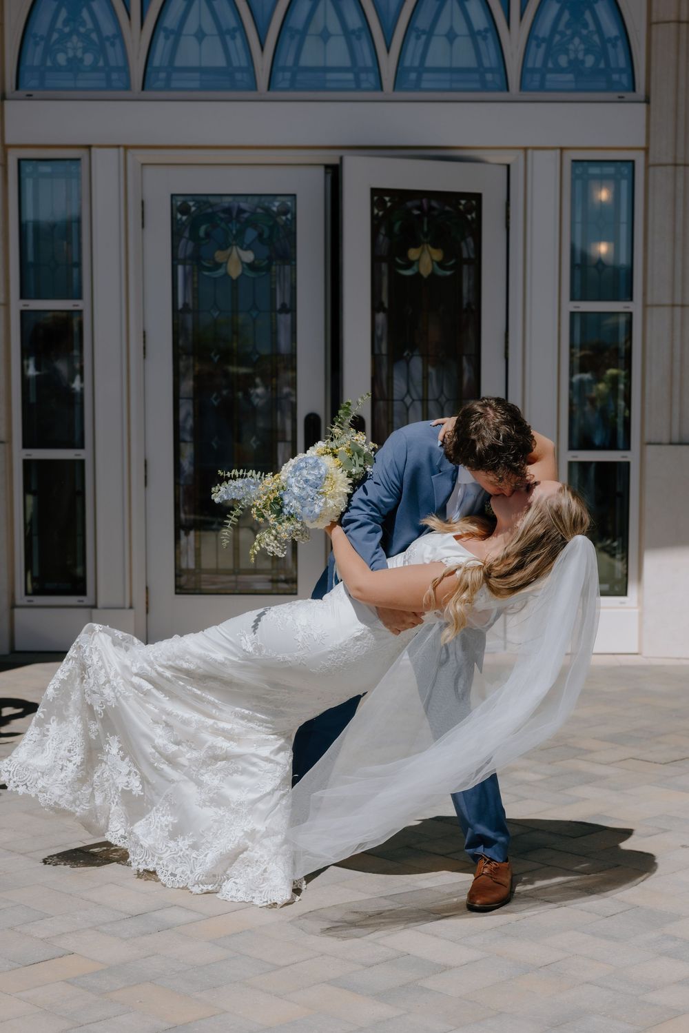 Groom dips bride in a romantic kiss outside elegant venue doors, bride in lace gown holding blue hydrangea bouquet.