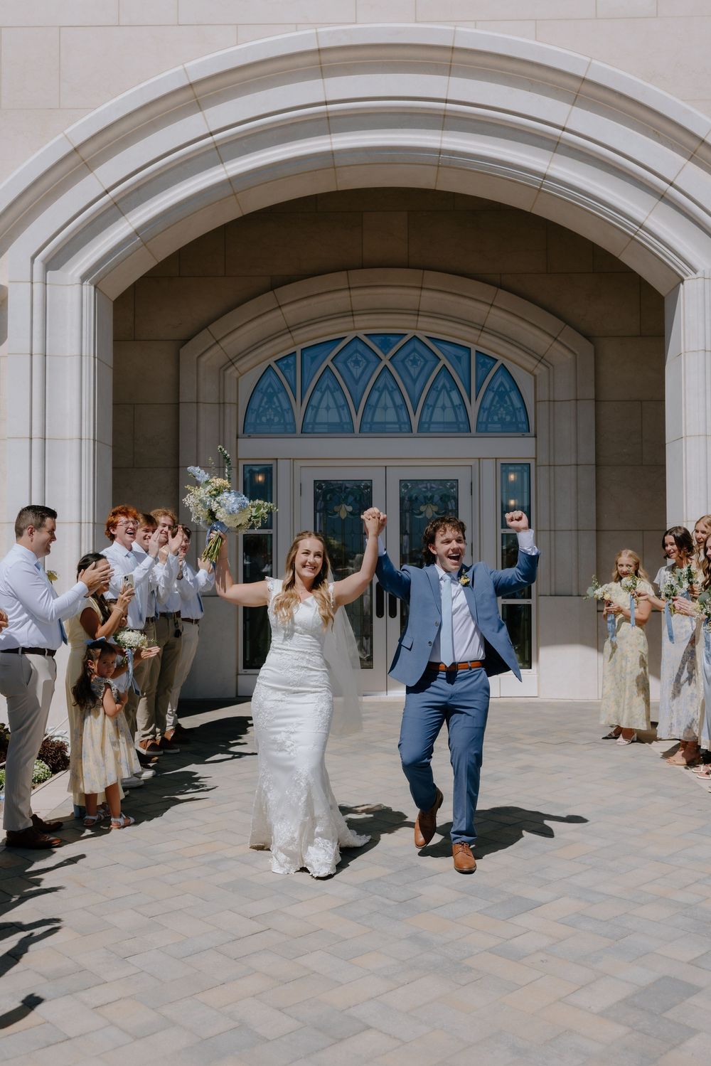 Joyful newlyweds exit a grand church arch, raising hands in celebration, surrounded by cheering guests in wedding attire.