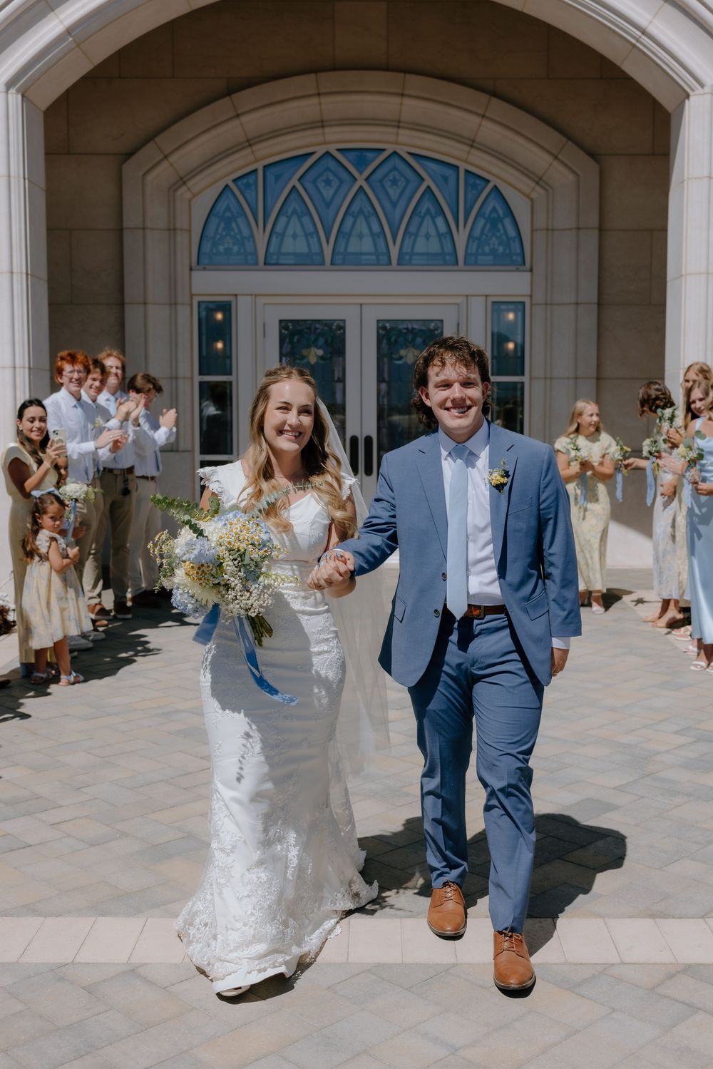 Joyful newlyweds exit church hand-in-hand in blue suit and lace gown, greeted by cheering wedding guests.