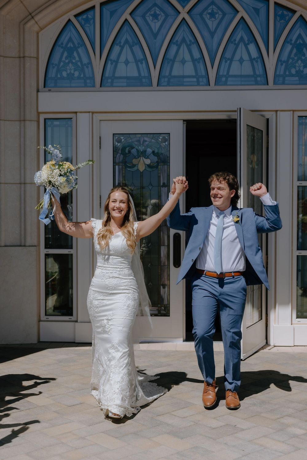 Joyful newlyweds celebrate outside church doors, bride in white gown holding bouquet, groom in blue suit raising fists.