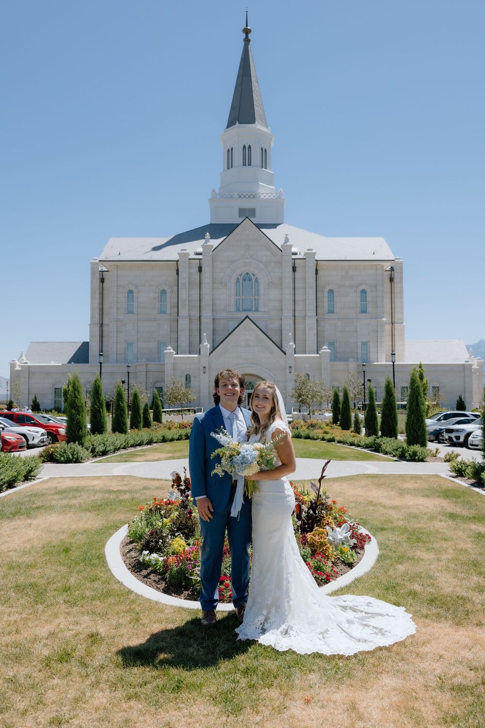Newlyweds standing together before a white LDS temple with a tall steeple and manicured gardens.