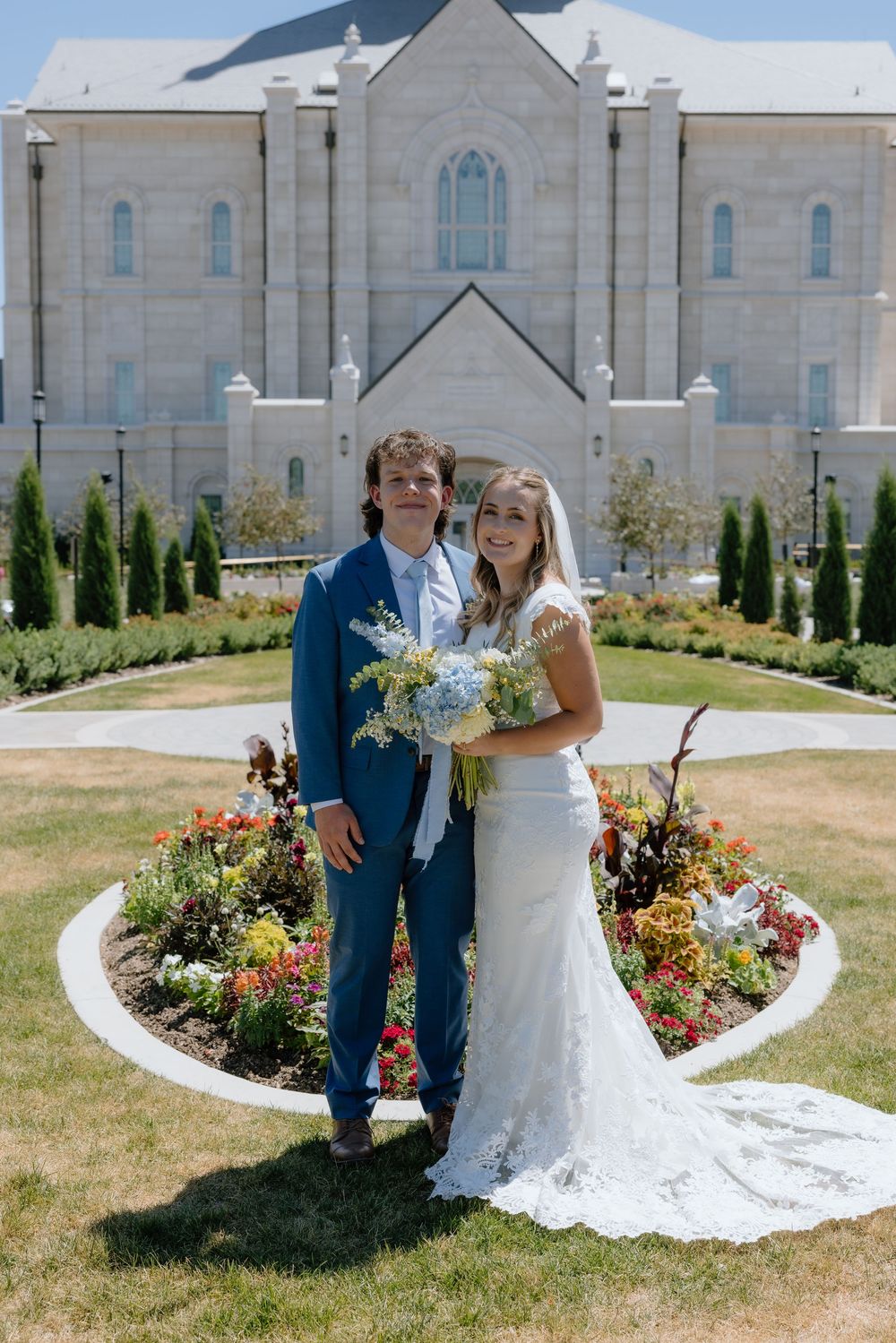 Bride and groom posing in front of an LDS temple garden with colorful flowers on their wedding day.