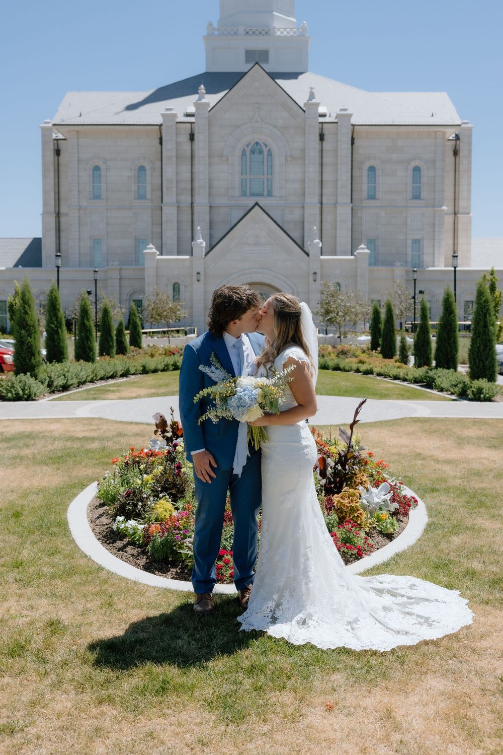 Couple sharing a kiss in front of a grand LDS temple surrounded by vibrant flower gardens.