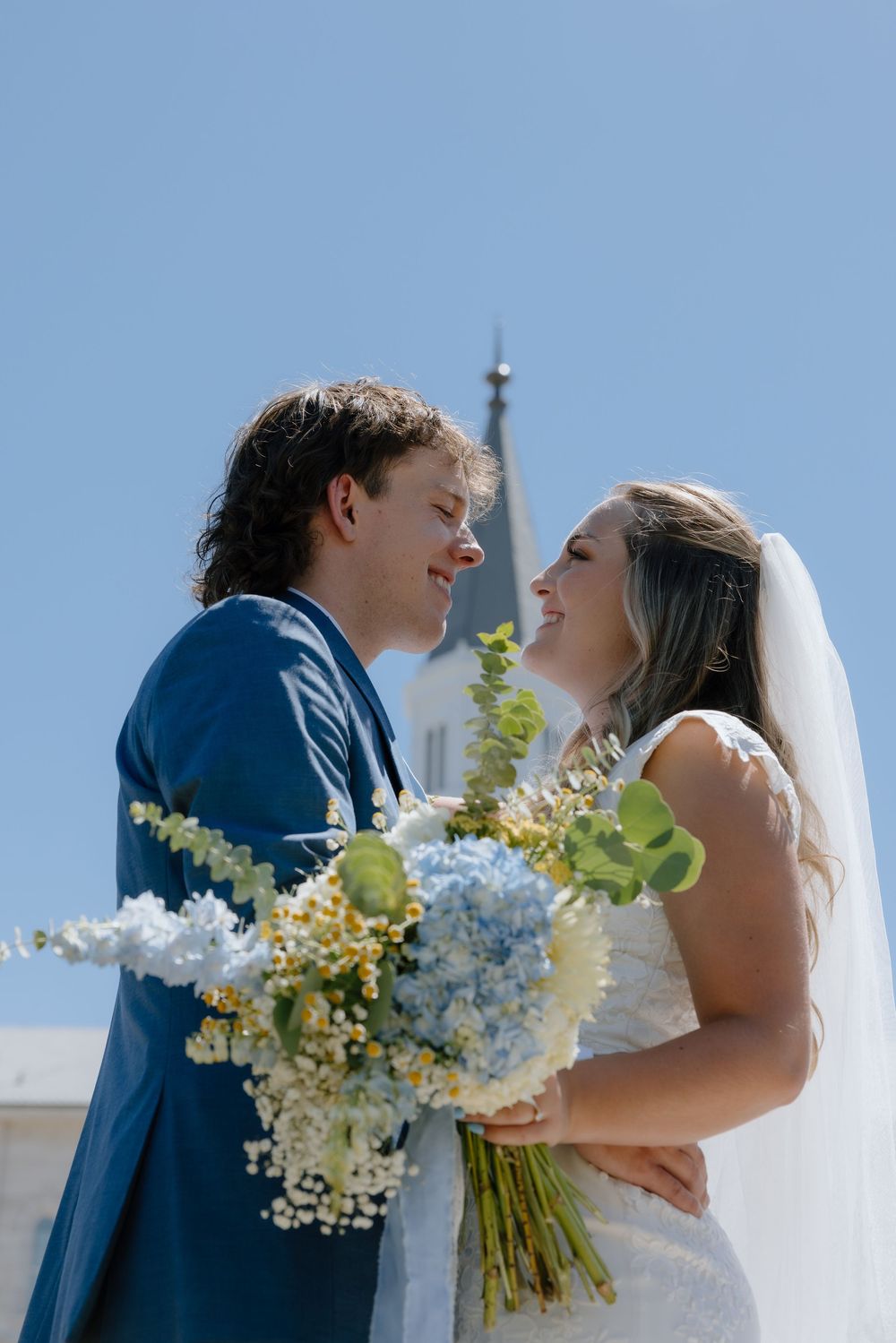 Bride and groom gazing at each other smiling, holding blue hydrangea bouquet under clear blue sky near church steeple.