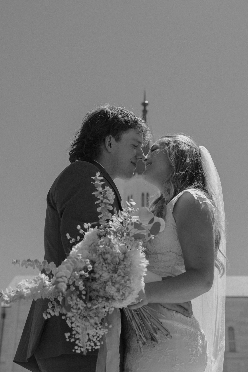 Black and white photo of bride and groom sharing a romantic kiss with a temple spire in background.