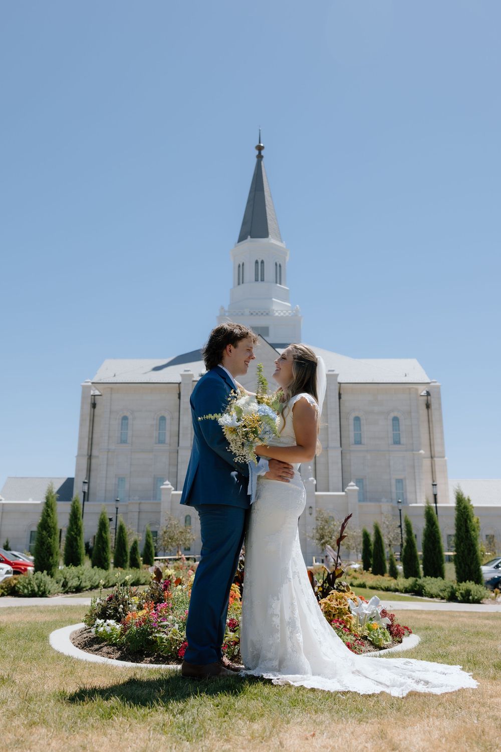 Bride and groom pose together in front of white LDS temple on sunny wedding day.