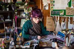 Woman with purple hair and glasses reads at a cluttered creative workspace surrounded by art supplies and handwritten notes.