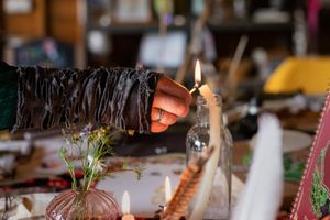 Hand in dark fringed sleeve lighting a candle on a mystical altar with crystals, feathers, and wildflowers.