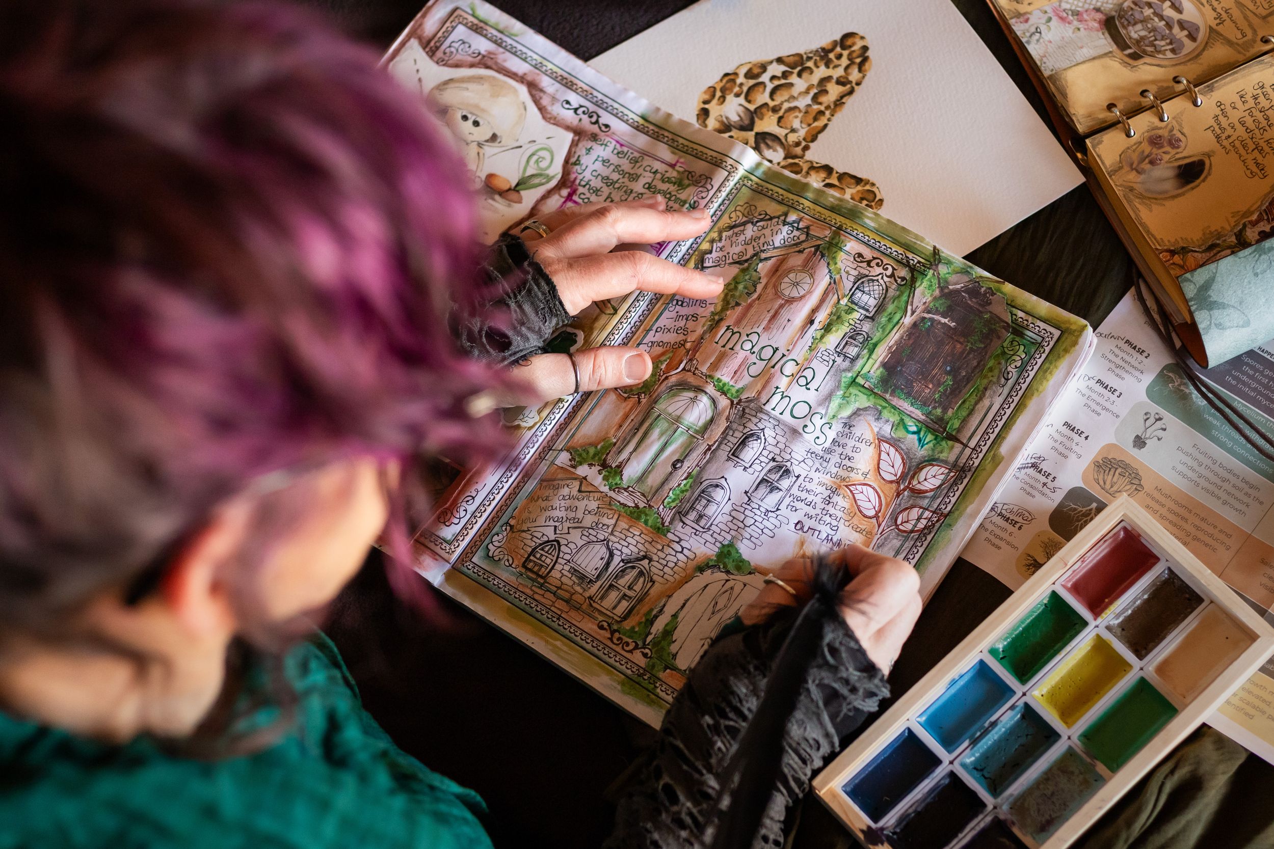 Artist with pink hair reviews a detailed illustrated sketchbook at a desk with watercolor paints nearby.
