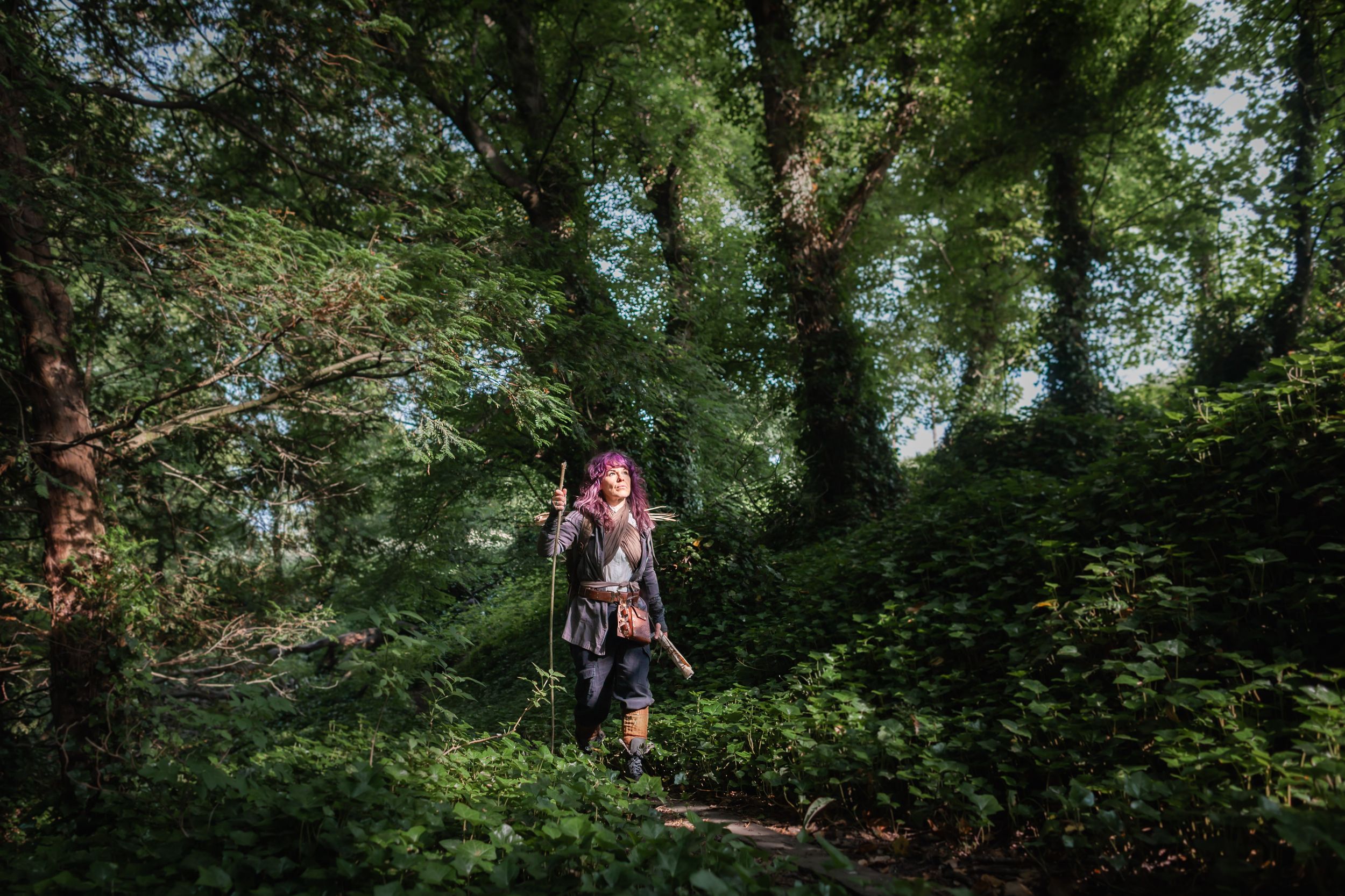 A costumed warrior with a staff walks through a lush green forest on a sunny day.