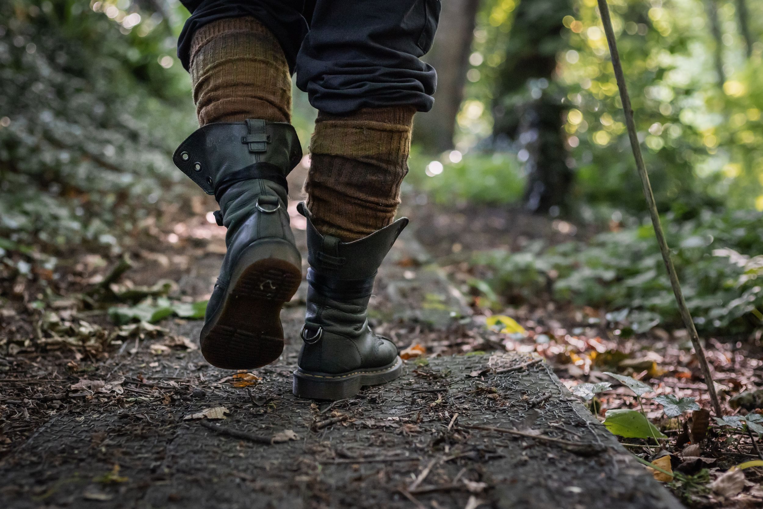Hiker walking on a forest trail wearing green boots and wool socks, holding a trekking pole among fallen leaves.