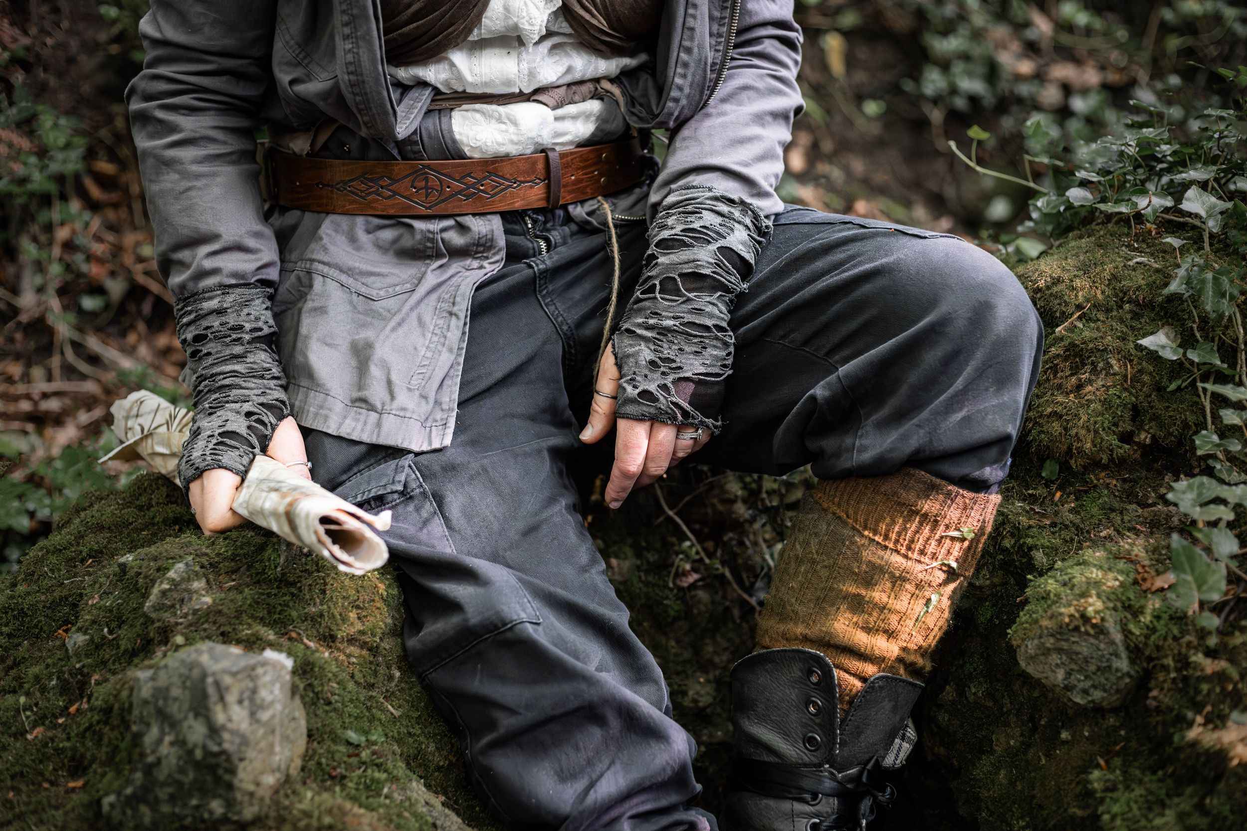 Medieval fantasy cosplay costume detail showing gray tunic, leather belt, dark pants, and boots on mossy rocks.