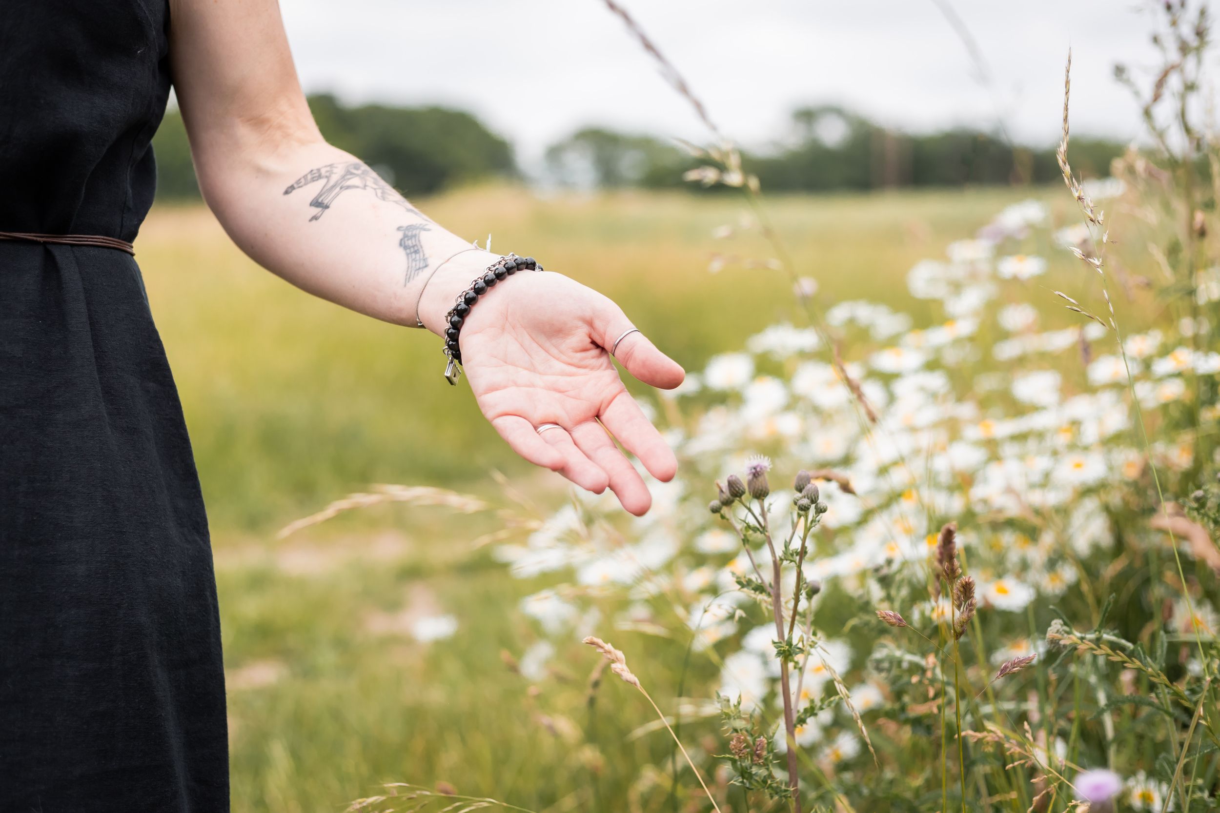 A tattooed hand reaches out over wildflowers in a meadow, wearing a beaded bracelet against a black sleeve.