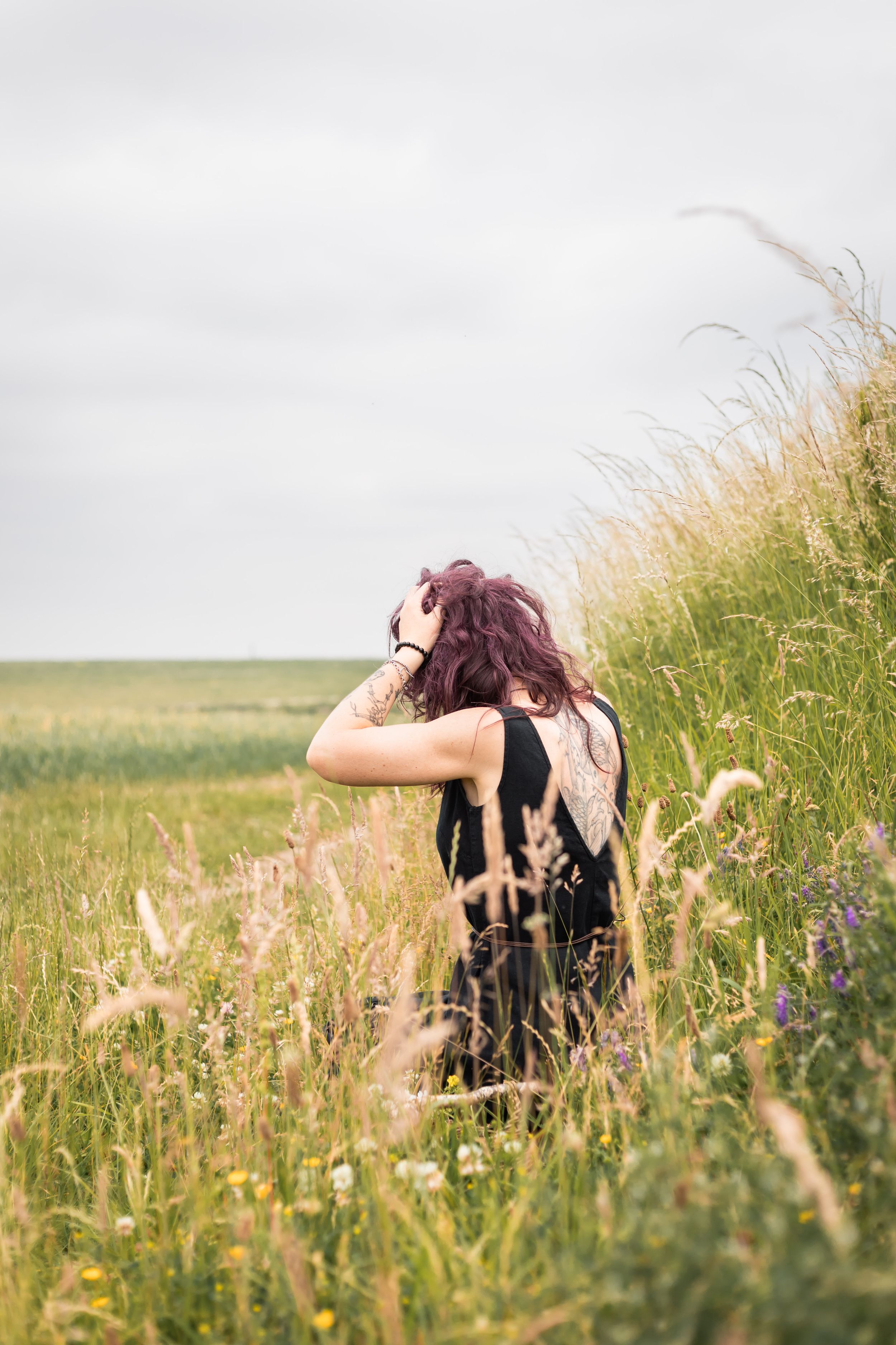 A woman with purple hair in a black dress stands in a wildflower field, facing away, touching her hair on a cloudy day.