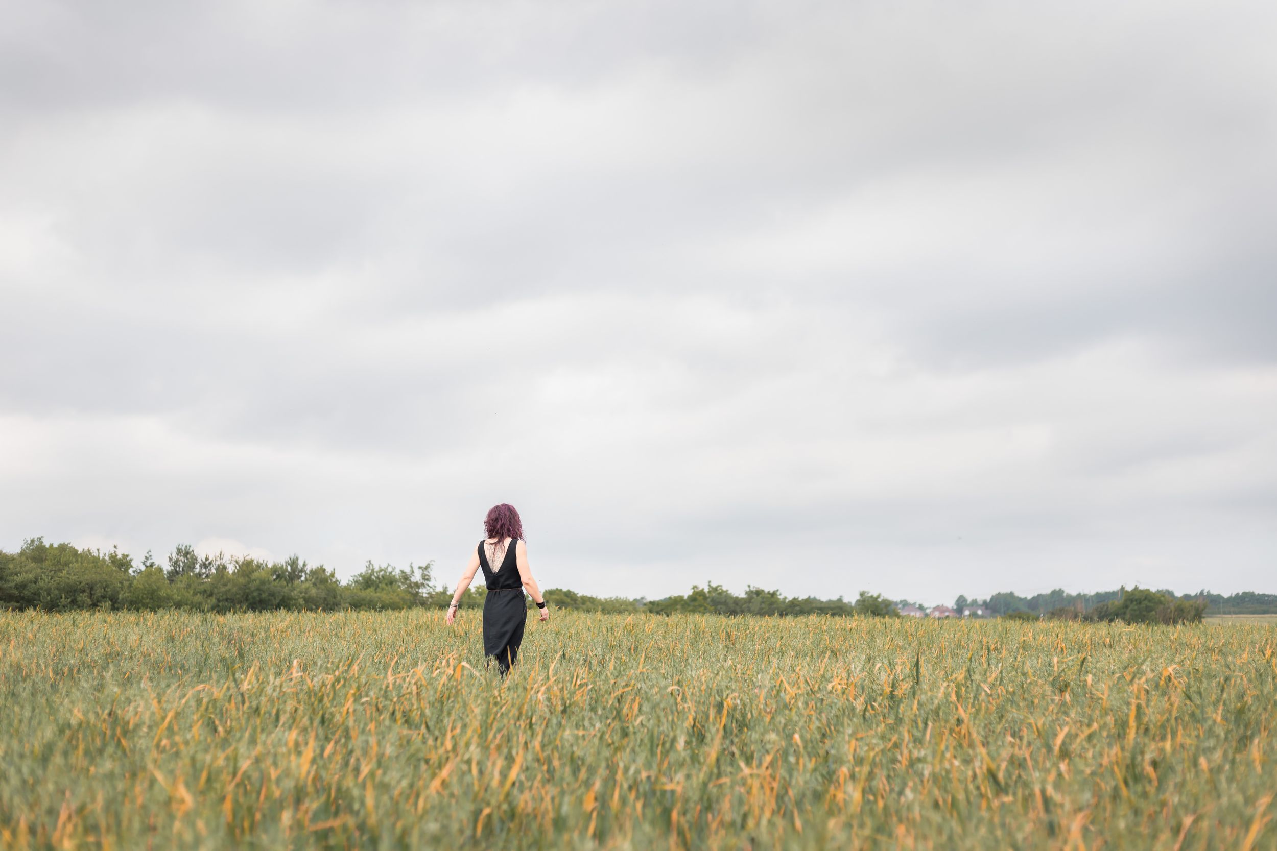 A woman in a black dress and red hat stands alone in a vast green field under a cloudy sky.