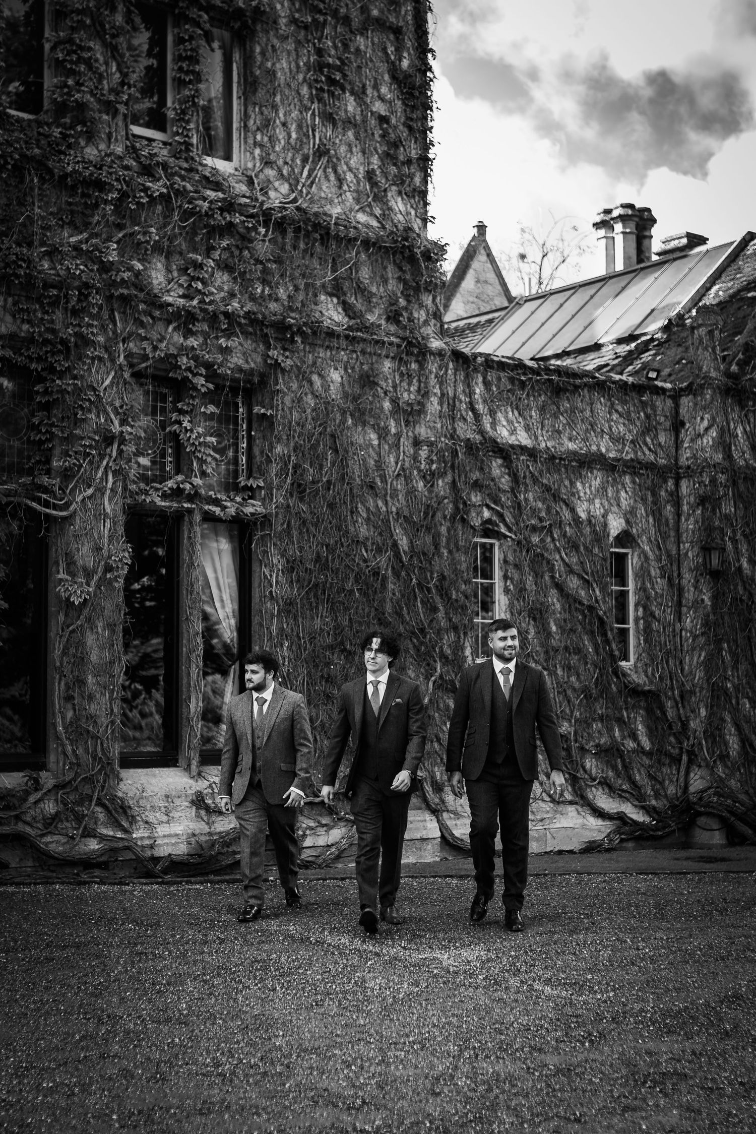 Three men in suits and vests walk together across a lawn in front of an ivy-covered stone manor in a black and white photo.