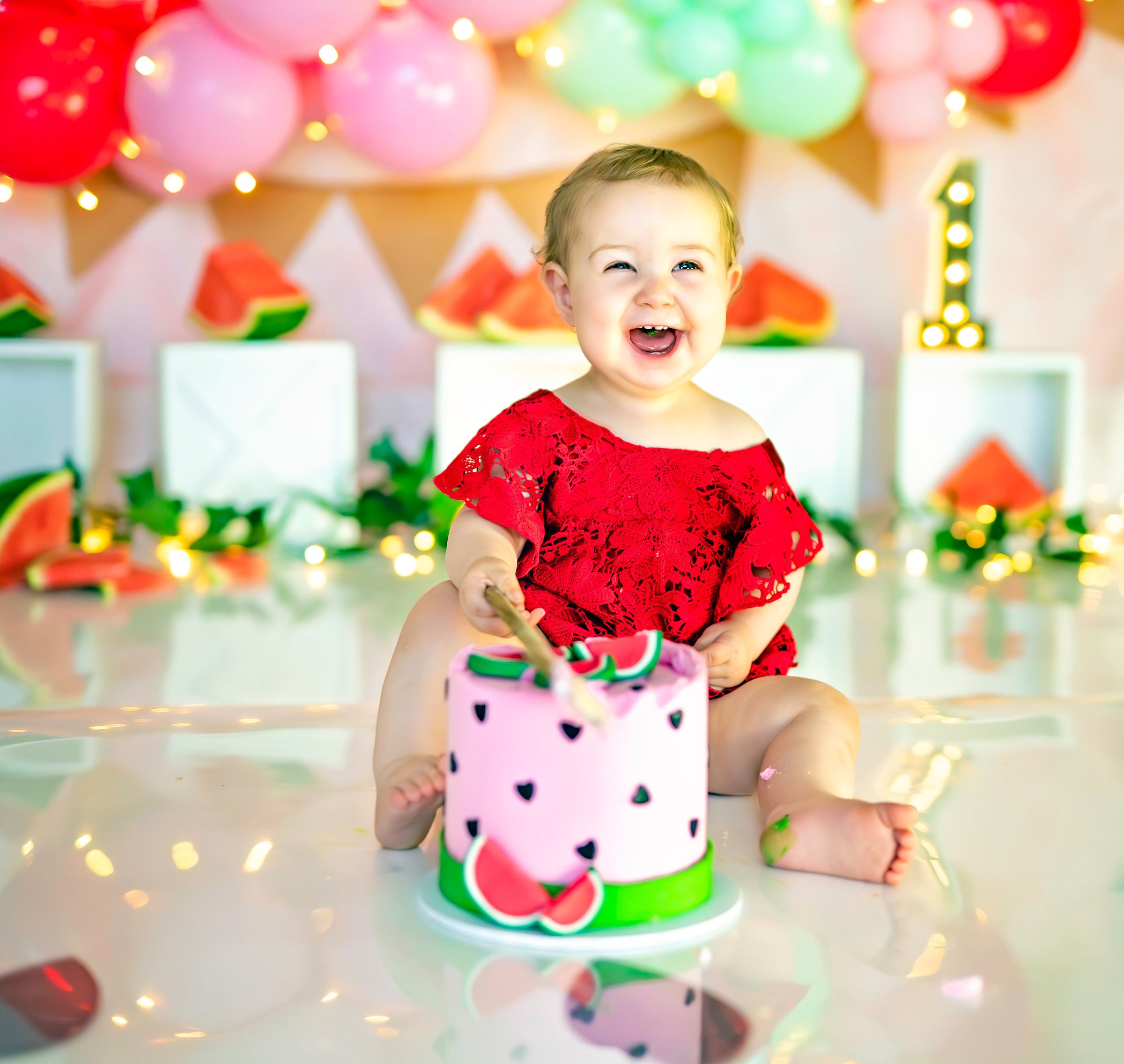 Baby girl in red sequin outfit smashes watermelon-themed first birthday cake surrounded by colorful balloons.