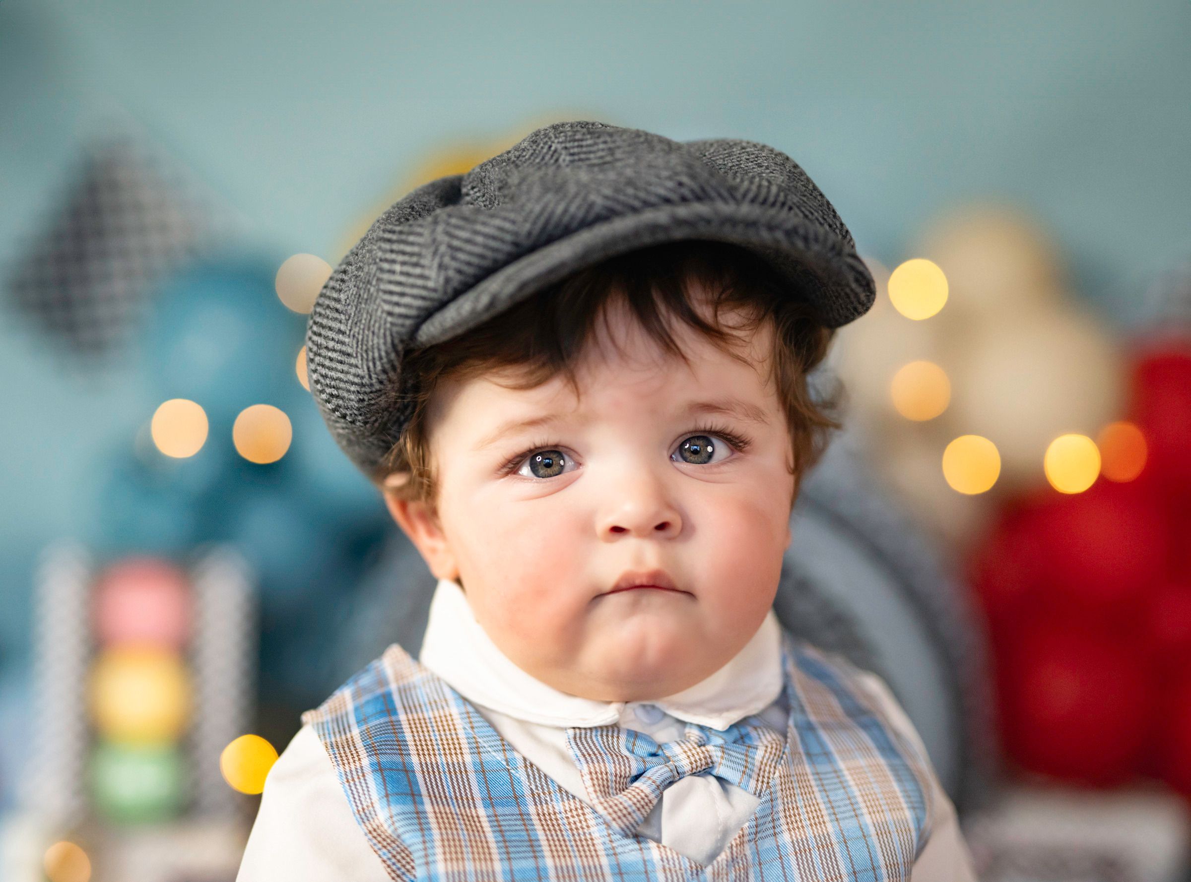 Adorable baby boy wearing a gray newsboy cap and plaid shirt with bow tie, posed against a bokeh light background.