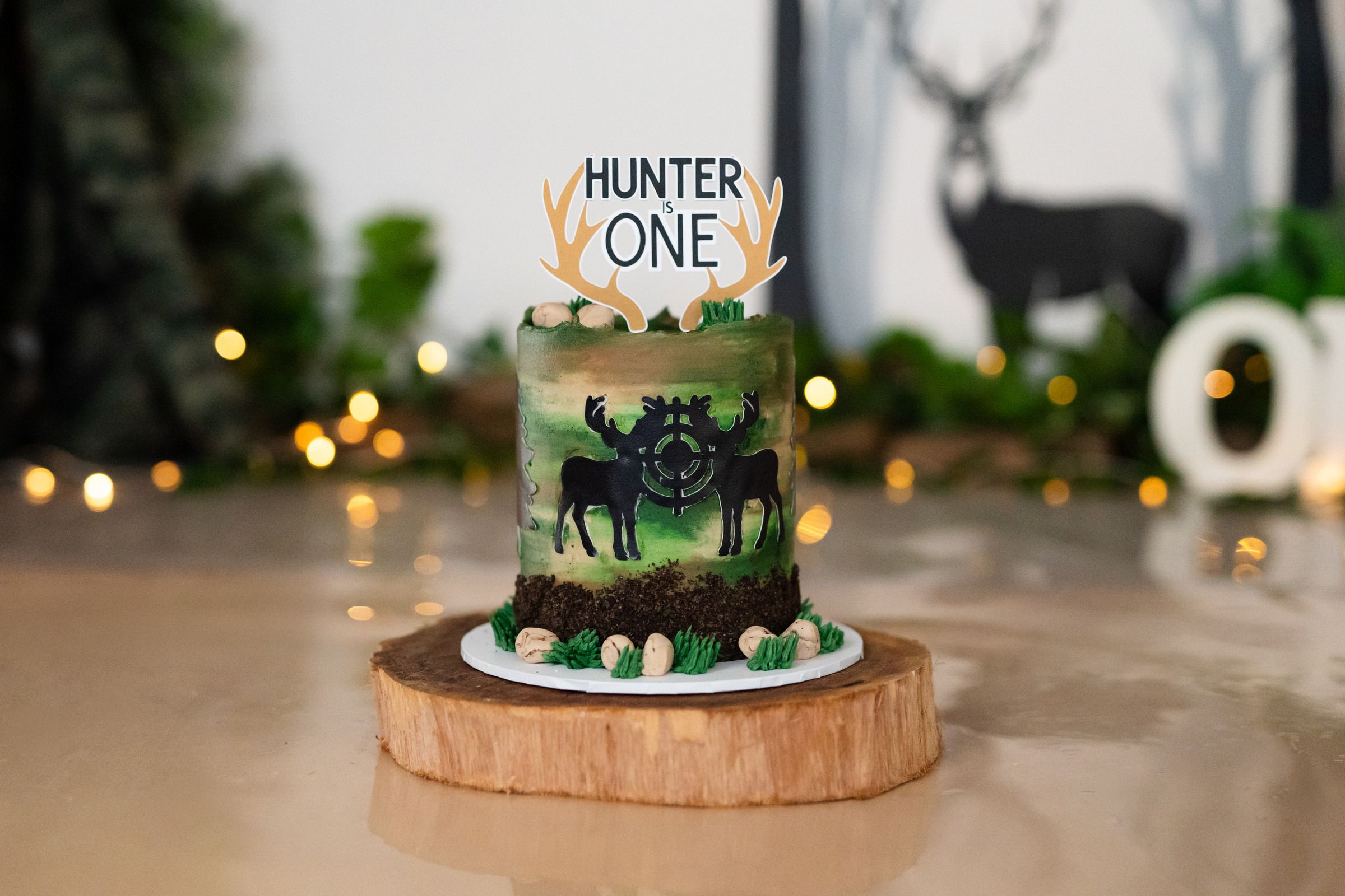 Hunter One first birthday cake with deer silhouette and antler topper on a wooden stand.
