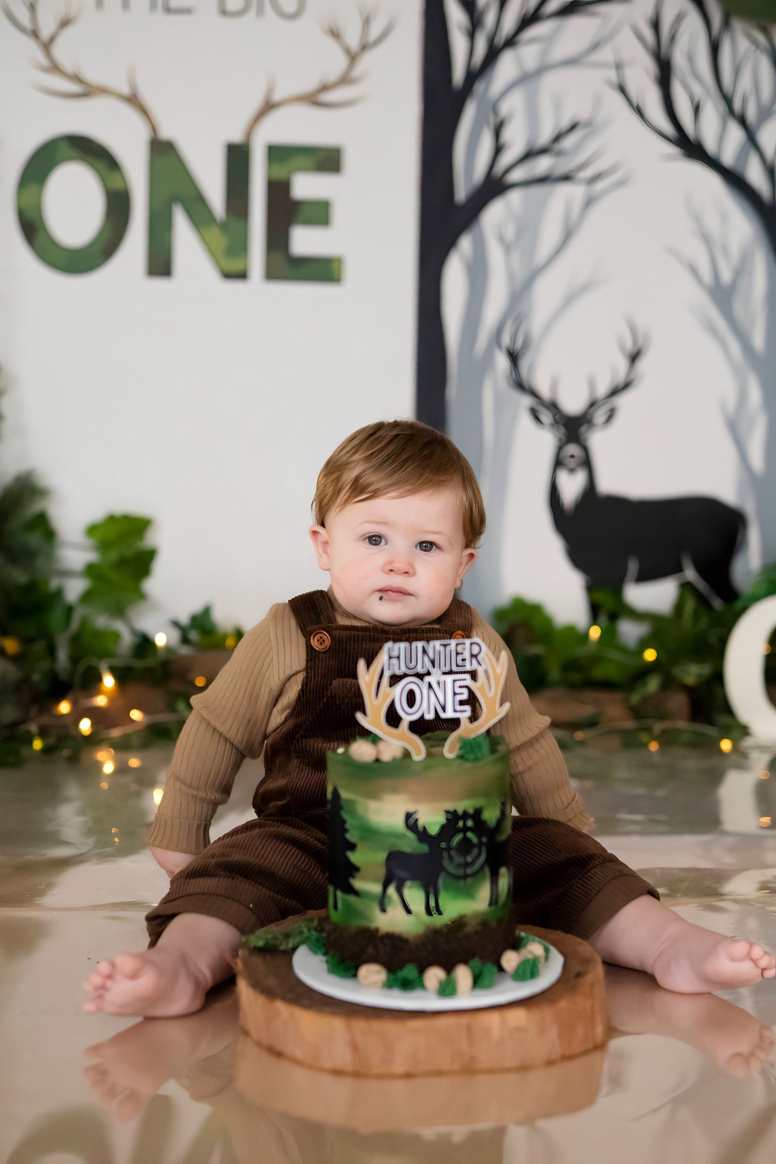 Baby boy in brown overalls sits before a hunting-themed first birthday cake topper reading Hunter One.