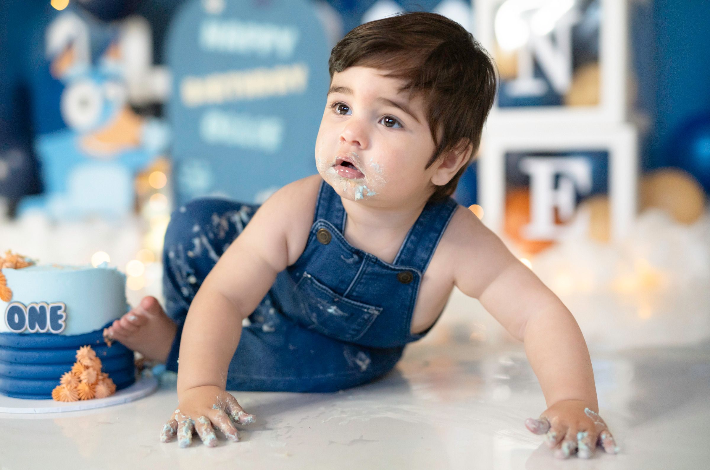 Baby boy in denim overalls celebrating first birthday smash cake session.
