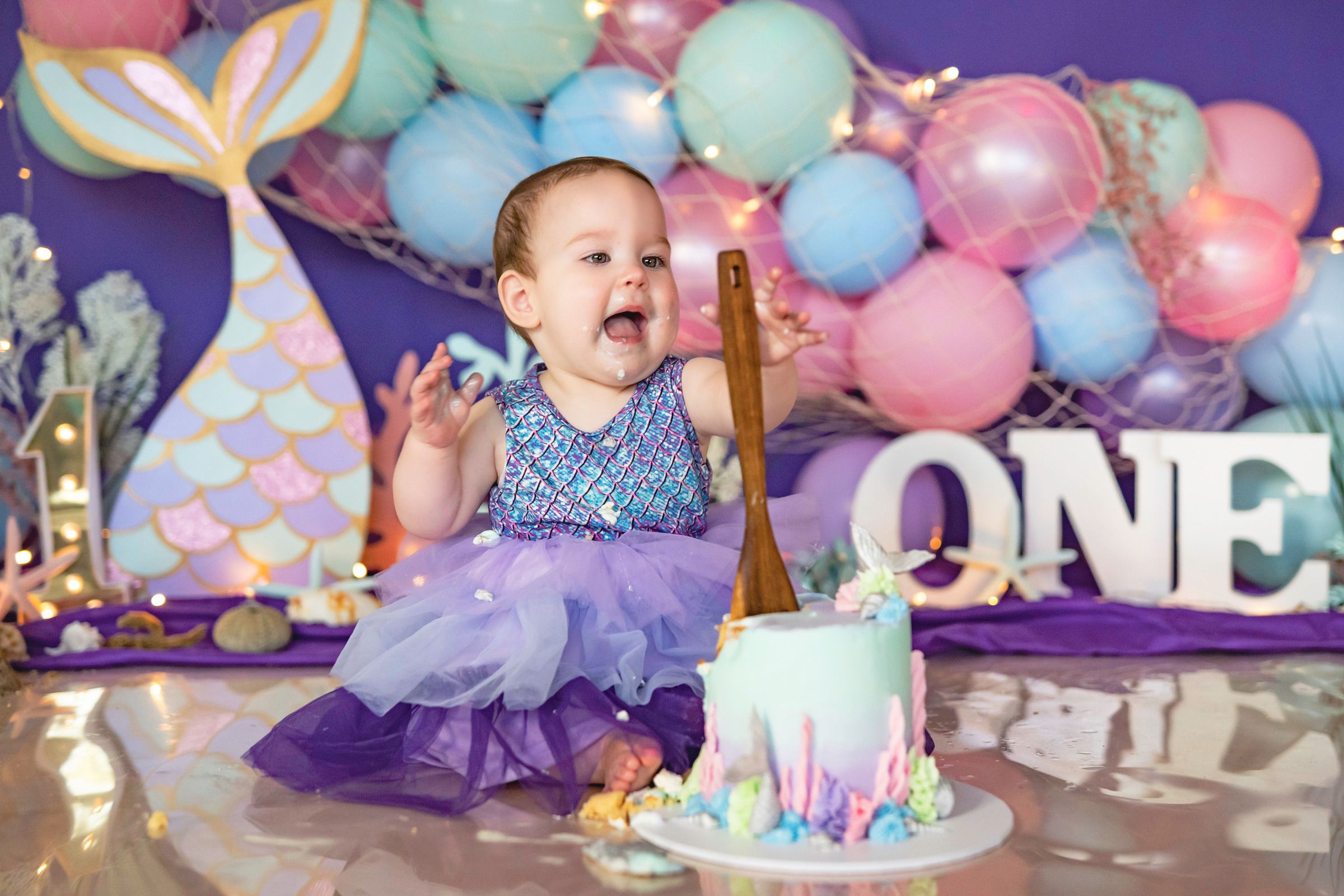 Baby girl in purple tutu celebrates first birthday with mermaid cake smash, balloons, and ONE sign backdrop.