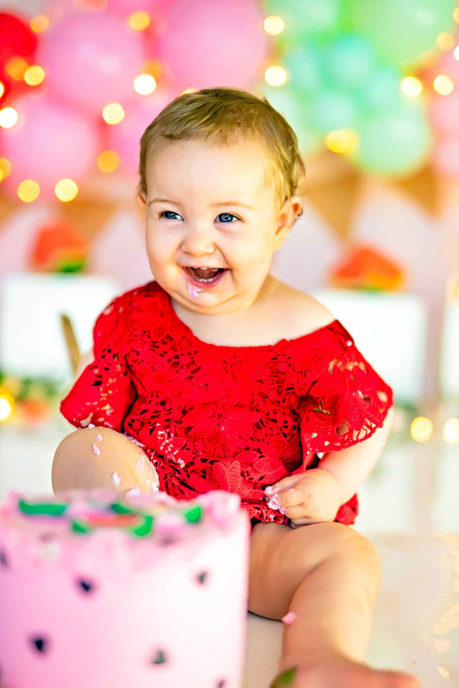 Happy baby girl in red lace dress smiling at first birthday cake smash with colorful balloon backdrop.