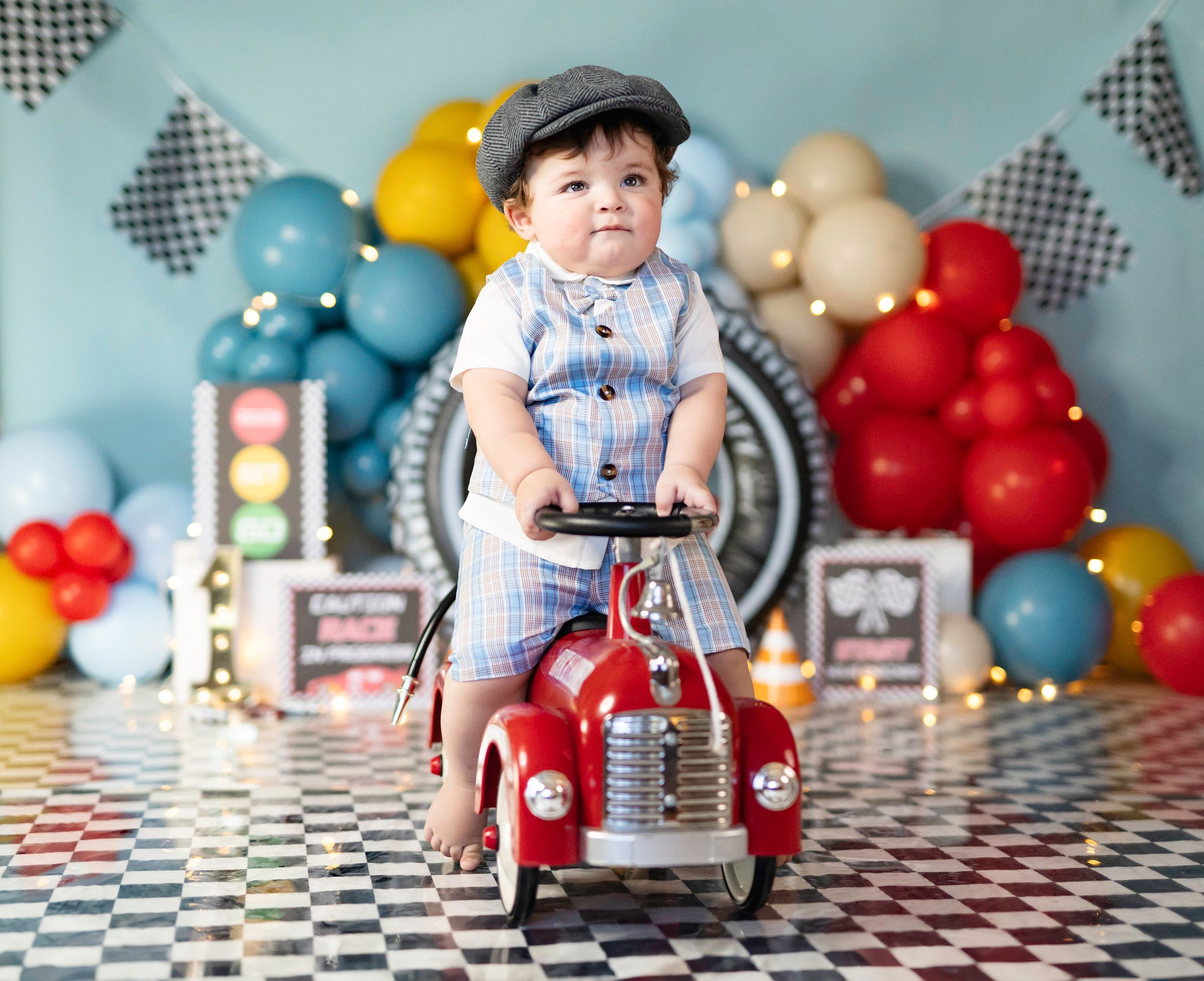 Cute baby boy in suspenders sits on a red toy car surrounded by colorful balloons for a race car birthday photo.