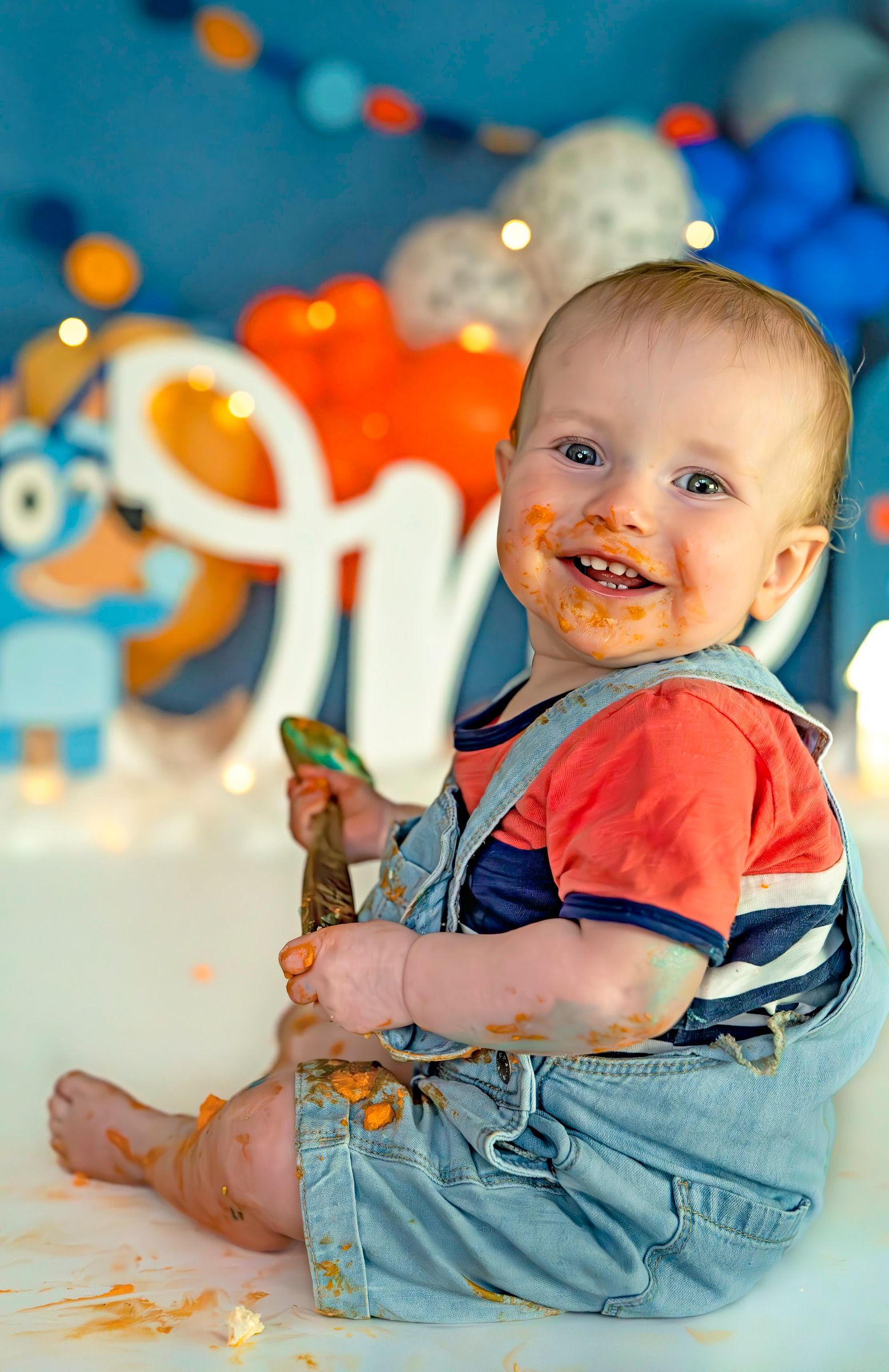Smiling baby boy in denim overalls enjoys a messy cake smash with orange frosting at a colorful first birthday party.