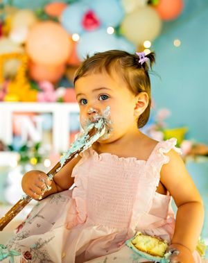 Baby girl in pink dress with cake smash frosting on face, holding cake topper, colorful balloon backdrop.