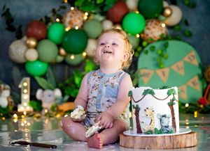 Baby smiling at first birthday cake smash with jungle theme decorations and green balloon garland backdrop.