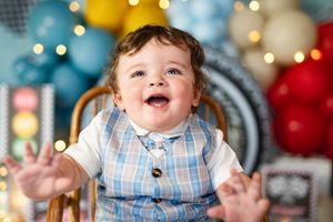 Happy baby boy in plaid vest smiles joyfully at first birthday party with colorful balloons.