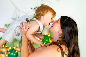 A mother joyfully lifts her laughing baby girl near a Christmas tree with bokeh lights in the background.
