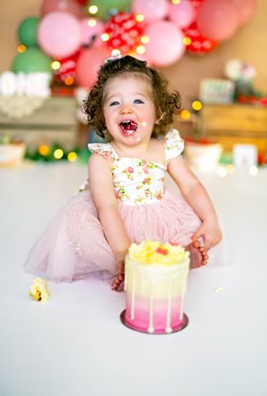 Toddler girl in pink tutu dress laughing joyfully at her first birthday cake smash with colorful balloon backdrop.