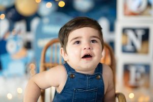 A baby boy in denim overalls sits in a high chair, looking up with wide eyes at a blue birthday party backdrop.