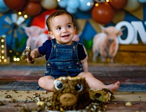 Baby boy in denim overalls smiling during a farm-themed first birthday cake smash photo session.